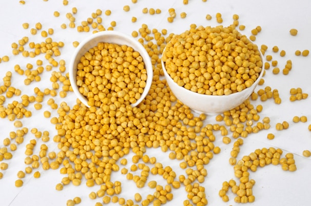 Several chickpeas spilled onto a flat surface. Two white bowls, filled with chickpeas, are placed nearby, with some of the legumes scattered around them.