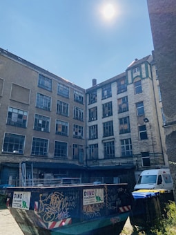 A large graffiti-covered dumpster is situated in an urban courtyard surrounded by tall, worn buildings with many windows. The sunlight is bright, casting harsh shadows on the scene. A white van and several trash bins are also visible in the area. The architecture appears aged and industrial.