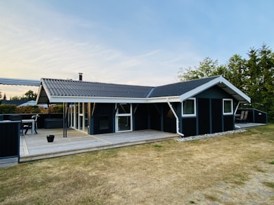 A modern, single-story house with dark exterior walls and a sloping roof, positioned on a wooden deck. It is surrounded by a neatly trimmed lawn and trees. The house features large windows and a chimney. Outdoor seating and a table are placed on the deck.