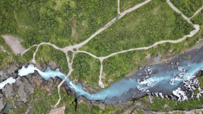 An aerial view captures a turquoise river snaking through rugged terrain with pathways traversing lush greenery. The river creates a series of curves and small waterfalls as it flows between patches of dense vegetation. The pathways suggest a walking trail often used by hikers or tourists.