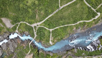 An aerial view captures a turquoise river snaking through rugged terrain with pathways traversing lush greenery. The river creates a series of curves and small waterfalls as it flows between patches of dense vegetation. The pathways suggest a walking trail often used by hikers or tourists.