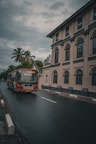 A red and white bus labeled 'Welcome' travels on a wet road next to a beige building with arched windows and columns. Palm trees and lush greenery can be seen in the background under a cloudy, overcast sky.