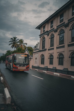 A red and white bus labeled 'Welcome' travels on a wet road next to a beige building with arched windows and columns. Palm trees and lush greenery can be seen in the background under a cloudy, overcast sky.