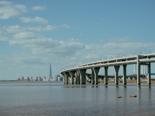 A modern bridge extends across a wide body of water with a cityscape visible in the distance. A tall skyscraper stands prominently on the horizon under a blue sky with scattered clouds.