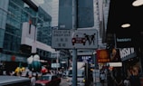 A street scene in a bustling city center with a focus on a signpost. The sign reads 'Pick Up & Drop Off' in both English and Chinese, with an image of a taxi and two people. The background features modern glass buildings, various shops, and people walking. Brightly colored sculptures resembling cartoon characters add a playful element to the scene.