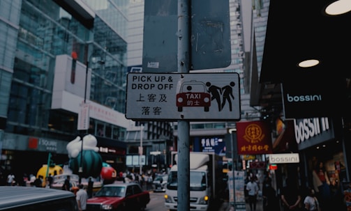 A street scene in a bustling city center with a focus on a signpost. The sign reads 'Pick Up & Drop Off' in both English and Chinese, with an image of a taxi and two people. The background features modern glass buildings, various shops, and people walking. Brightly colored sculptures resembling cartoon characters add a playful element to the scene.