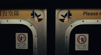 A close-up view of two metal doors, likely from a subway or train car. Each door has warning signs in both English and another language, advising passengers not to block or attempt to board when the doors are closing. The signs include simple graphics depicting prohibited actions.