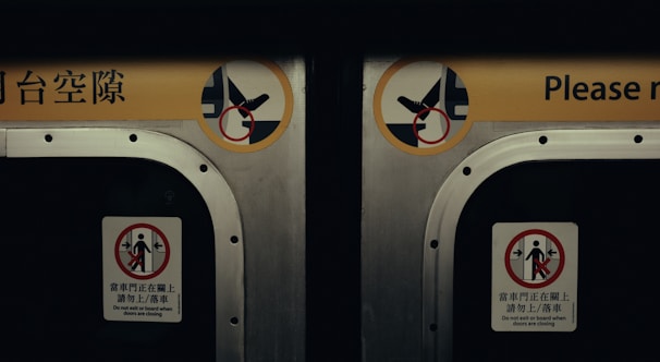 A close-up view of two metal doors, likely from a subway or train car. Each door has warning signs in both English and another language, advising passengers not to block or attempt to board when the doors are closing. The signs include simple graphics depicting prohibited actions.