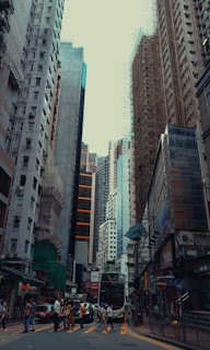 Tall buildings line a bustling city street with pedestrians crossing and a double-decker bus approaching. Scaffolding is visible on some of the structures, indicating ongoing construction. The urban setting is densely packed with a mix of modern and older architecture.