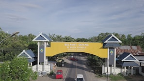 A large, welcoming yellow archway spans across a road, with the text 'SELAMAT DATANG DI BUTTA TURATEA' and 'KABUPATEN JENEPONTO' visible on it. The arch is supported by two white pillars with dark blue panels and roof-like structures on top. Surrounding the area, there are green trees, residential structures with blue roofs, and the landscape extends under a blue sky with some clouds.