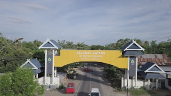 A large, welcoming yellow archway spans across a road, with the text 'SELAMAT DATANG DI BUTTA TURATEA' and 'KABUPATEN JENEPONTO' visible on it. The arch is supported by two white pillars with dark blue panels and roof-like structures on top. Surrounding the area, there are green trees, residential structures with blue roofs, and the landscape extends under a blue sky with some clouds.