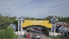 A large, welcoming yellow archway spans across a road, with the text 'SELAMAT DATANG DI BUTTA TURATEA' and 'KABUPATEN JENEPONTO' visible on it. The arch is supported by two white pillars with dark blue panels and roof-like structures on top. Surrounding the area, there are green trees, residential structures with blue roofs, and the landscape extends under a blue sky with some clouds.