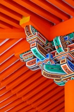 Intricately painted wooden architectural elements featuring vibrant colors, intricate patterns, and traditional carving. The structure is primarily composed of parallel wooden beams painted in a striking orange, complemented by geometric and floral motifs in green, blue, red, and black.