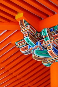 Intricately painted wooden architectural elements featuring vibrant colors, intricate patterns, and traditional carving. The structure is primarily composed of parallel wooden beams painted in a striking orange, complemented by geometric and floral motifs in green, blue, red, and black.