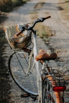 A bicycle is parked on a gravel path surrounded by grass. The bike features a brown leather seat and handles, with a wicker basket attached to the front, holding a rolled-up mat. Sunlight casts a warm glow, highlighting the rustic and serene setting.