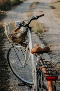 A bicycle is parked on a gravel path surrounded by grass. The bike features a brown leather seat and handles, with a wicker basket attached to the front, holding a rolled-up mat. Sunlight casts a warm glow, highlighting the rustic and serene setting.