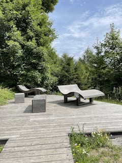 A wooden deck is surrounded by lush green trees in a natural outdoor setting. On the deck, there are two modern, curved wooden lounge chairs and two cuboid benches positioned neatly. The sky is partially visible, dotted with light clouds.