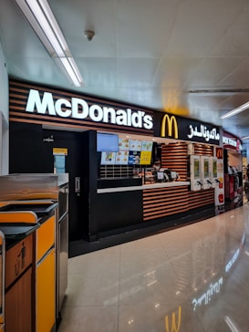 A McDonald's restaurant counter is situated in a mall or airport environment. The design features a modern look with black and wooden stripes. A bright McDonald's sign is displayed prominently. Trash bins are visible to the side, and digital menus are positioned above the counter.