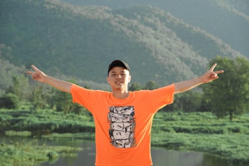 A person wearing an orange shirt with anime print and a black cap stands with arms outstretched in a lush green landscape, with mountains in the background and a small body of water nearby.