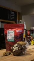 A package of Eurocafe coffee is placed on a wooden surface along with a clear plastic bag containing various cookies. The background is a cafe setting with a chalkboard menu and some shelves.