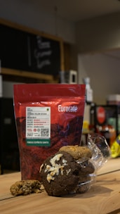 A package of Eurocafe coffee is placed on a wooden surface along with a clear plastic bag containing various cookies. The background is a cafe setting with a chalkboard menu and some shelves.