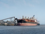 A large cargo ship is docked at an industrial port. The vessel is surrounded by cranes and conveyor belts for loading and unloading materials. The water is calm, and the sky is clear with a hint of blue.