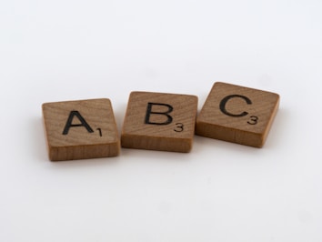 Three wooden Scrabble tiles are arranged in a subtle arc on a plain white background, each displaying a letter and a corresponding point value in black font. The letters are A, B, and C.