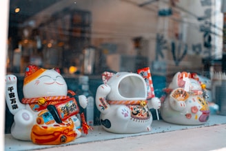 Three maneki-neko figurines are displayed in a row behind a glass window. Each figure has a raised paw, traditionally believed to bring good luck. The figurines are adorned with colorful details, including red and gold accents, decorative sashes, and various ornaments. The scene captures a shop-like setting with blurred background elements.