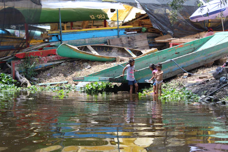Children playing by the lakeside with colorful boats and green trees in the background.
