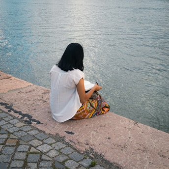 A person with long dark hair sits near a waterfront, writing or drawing in a book. They wear a white shirt and a colorful patterned skirt. The setting includes a stone pavement and calm water, creating a peaceful atmosphere.