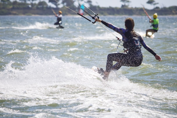 Three kite surfers navigate the waves on a sunny day, with one prominently in the foreground, skillfully maneuvering the board as the kite strings extend into the sky. The water splashes dynamically around them, and distant shoreline and trees are faintly visible in the background.