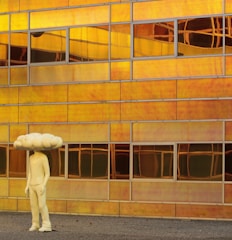 A sculpture resembling a human figure with a large cloud-shaped head stands in front of a modern building facade. The building is constructed with reflective panels that display a rich, warm, golden hue, creating a surreal and artistic backdrop. The reflective panels are rectangular and vary in their alignment and reflection due to the light and architecture.