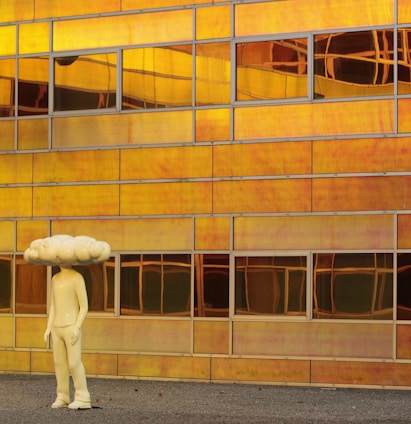 A sculpture resembling a human figure with a large cloud-shaped head stands in front of a modern building facade. The building is constructed with reflective panels that display a rich, warm, golden hue, creating a surreal and artistic backdrop. The reflective panels are rectangular and vary in their alignment and reflection due to the light and architecture.