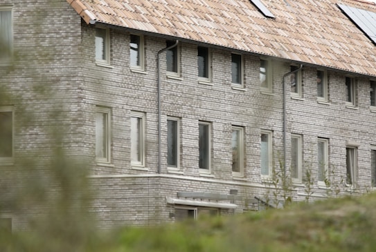 A multi-story brick building with a tiled roof is partially obscured by vegetation in the foreground. The structure features multiple windows and solar panels on the roof, suggesting an environmentally conscious design.