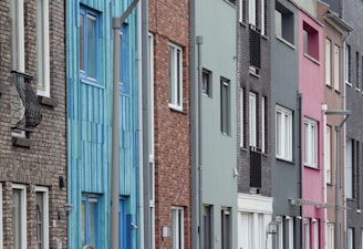 A row of modern, narrow urban houses with diverse facades, featuring a mix of materials and colors including brick, blue, gray, and pink tones. The windows are evenly spaced, and there are metal railings and street lamps visible.