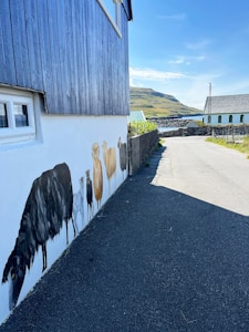 A rustic blue wooden building with a painted mural of sheep on a white wall. The building is adjacent to a narrow country road, surrounded by stone fences and lush green fields with rolling hills in the background under a bright blue sky.