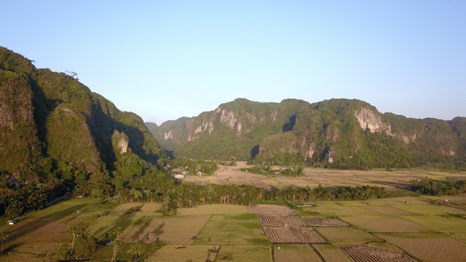 Expansive rural landscape featuring lush green fields interspersed with patches of brown farmland, bordered by towering, forested mountains in the background. The scene conveys a sense of tranquility, with clear blue skies above.