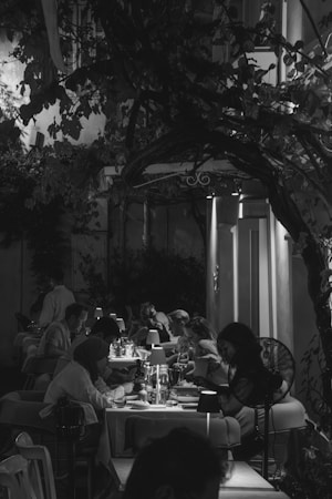A black and white photograph of a restaurant with several people dining at tables. The ambiance is intimate with soft lighting provided by small lamps on each table. Overhead, there are branches and leaves creating a natural canopy. The setting appears to be outdoors with a cozy and warm atmosphere.