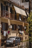 A residential building with balconies featuring metal railings and covered with retractable awnings. Air conditioning units are installed on the walls. Clothes are hanging to dry on a line outside the windows. In front of the building, a small car is parked. The scene is captured in bright, natural light, indicating a sunny day.
