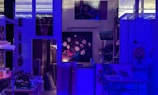 An art studio illuminated by blue lighting, creating a moody atmosphere. There are several paintings displayed on the walls, including a central one depicting a bouquet of flowers. Art supplies, such as brushes and paints, are organized on shelves and a table.