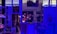 An art studio illuminated by blue lighting, creating a moody atmosphere. There are several paintings displayed on the walls, including a central one depicting a bouquet of flowers. Art supplies, such as brushes and paints, are organized on shelves and a table.