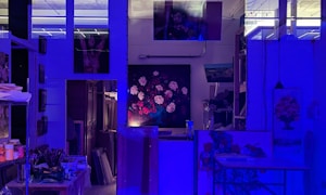 An art studio illuminated by blue lighting, creating a moody atmosphere. There are several paintings displayed on the walls, including a central one depicting a bouquet of flowers. Art supplies, such as brushes and paints, are organized on shelves and a table.