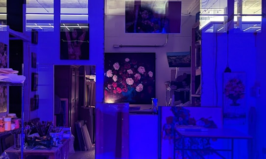 An art studio illuminated by blue lighting, creating a moody atmosphere. There are several paintings displayed on the walls, including a central one depicting a bouquet of flowers. Art supplies, such as brushes and paints, are organized on shelves and a table.