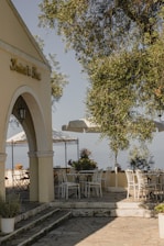 A relaxed outdoor patio setup featuring comfortable seating, light fabrics, and greenery under a clear blue sky.