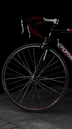 A close-up view of a bicycle's front wheel and handlebars. The bike features a red and black color scheme, with intricate details visible on the metal spokes and frame. The background is dark, emphasizing the bicycle's sleek design.