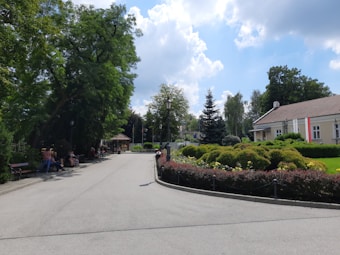 A peaceful park scene with a paved path bordered by benches and people sitting. There are lush green trees and manicured bushes with yellow flowers. To the right, a building with a red and white flag is visible. The sky is partly cloudy, creating a serene atmosphere.