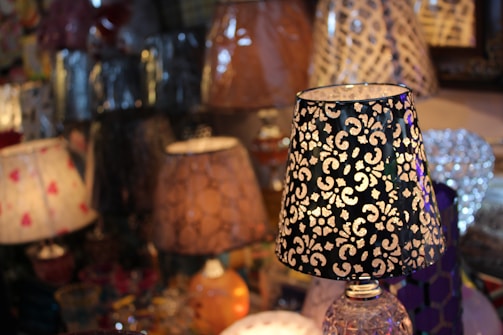 A collection of decorative table lamps with various ornate designs and patterns on their shades. The foreground features a lamp with a black shade adorned with intricate white floral motifs. The background is filled with numerous other lamps, each with unique colors and textures, creating a visually rich and warm atmosphere.