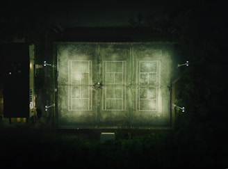 An aerial view of a tennis court complex taken at night, featuring four rectangular courts illuminated by floodlights. The surrounding area is dark with some foliage visible on one side and a building structure on the other. The courts have clear white lines marking the various sections.