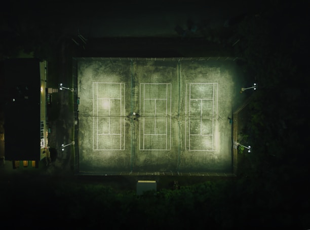An aerial view of a tennis court complex taken at night, featuring four rectangular courts illuminated by floodlights. The surrounding area is dark with some foliage visible on one side and a building structure on the other. The courts have clear white lines marking the various sections.