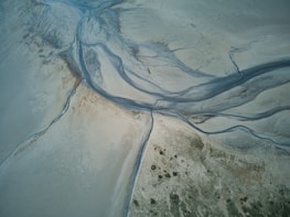 An aerial view of abstract natural patterns created by rivers and streams flowing through a sandy or muddy landscape. The scene features winding channels with shades of blue and gray merging into the surrounding beige and green textures.